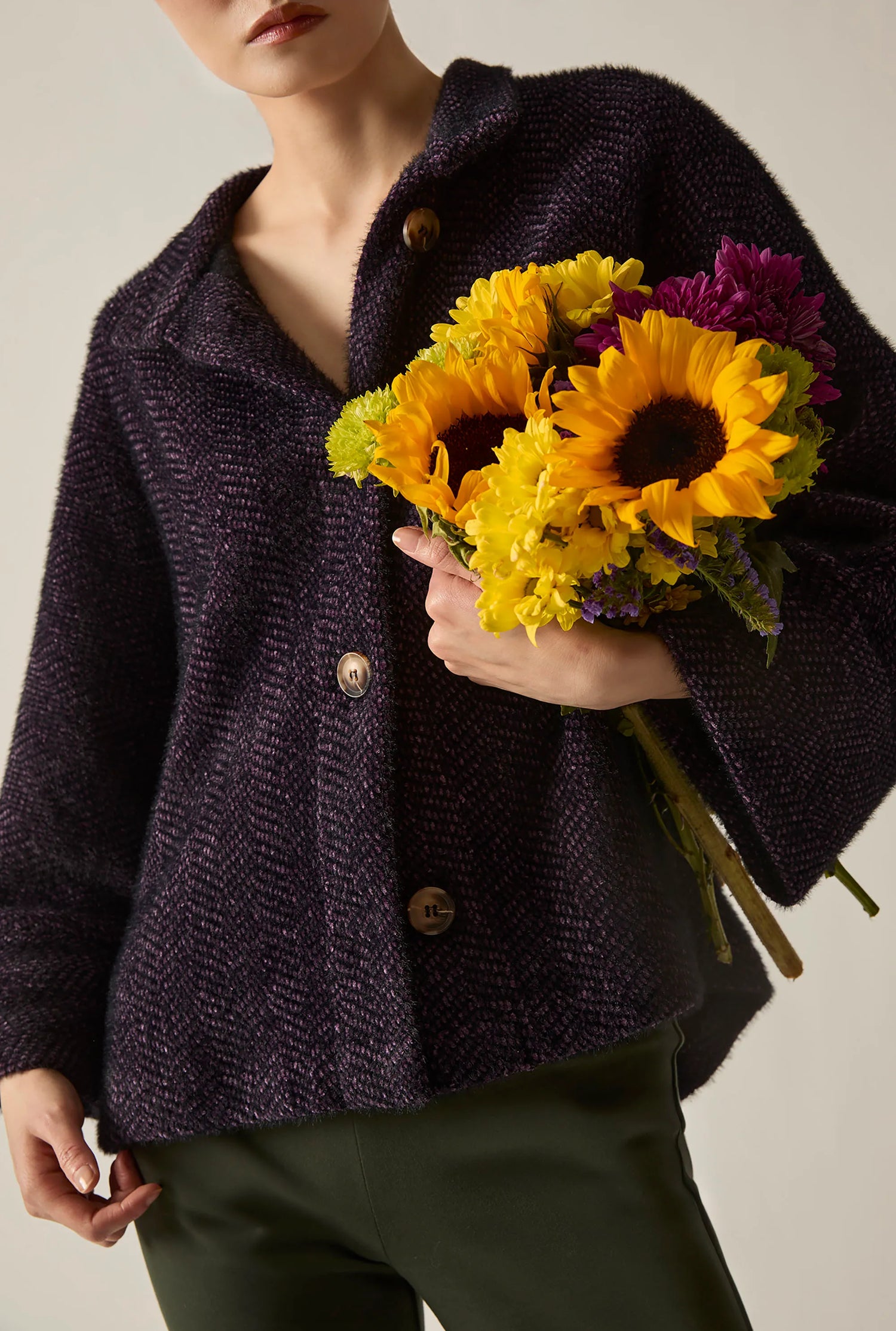 Person wearing a black and purple batwing jacket with olive trousers, holding a colourful bouquet of flowers.