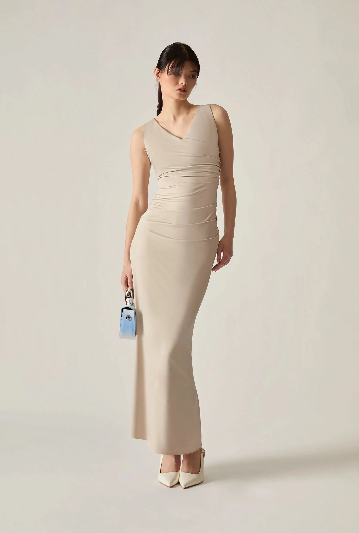 Full‑body shot of a woman wearing a beige dress, holding a small blue bag in her right hand against a soft beige background.