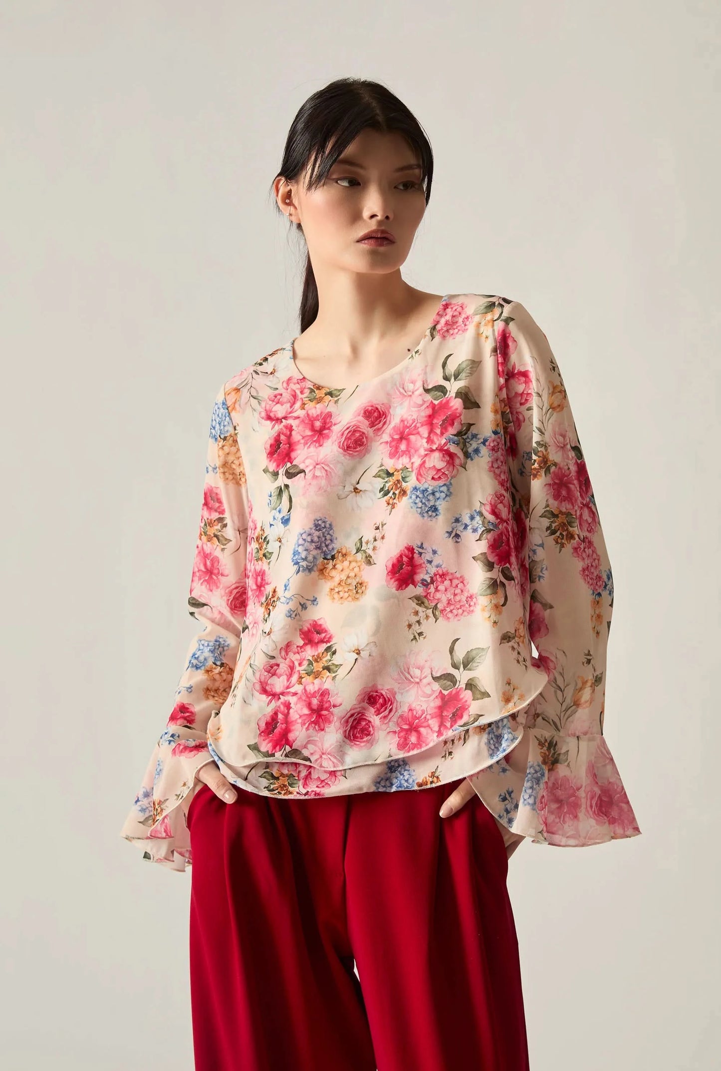 Woman wearing a floral blouse with red wide‑leg trousers, standing against a soft beige background.