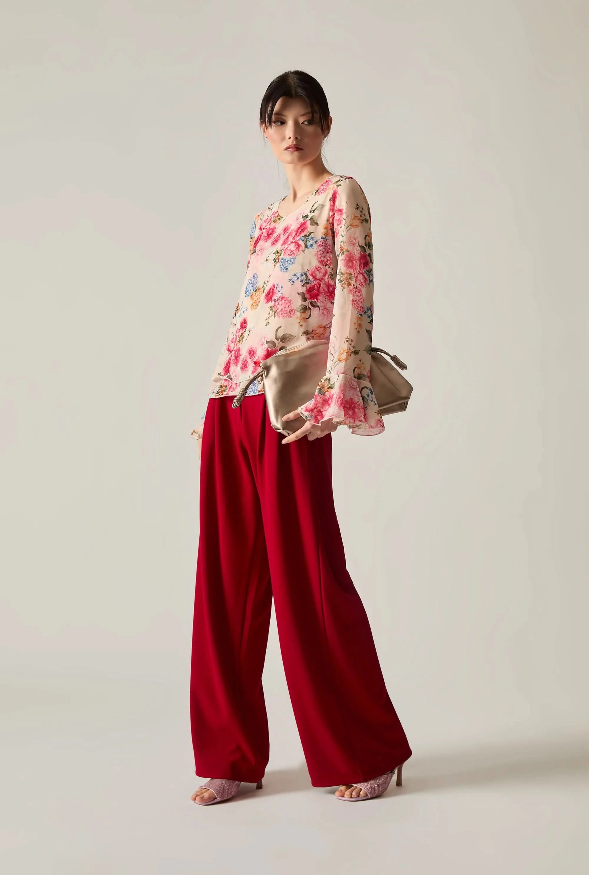Full‑body shot of a woman wearing a floral blouse with red wide‑leg trousers, holding a beige bag and wearing pink shoes against a soft beige background.