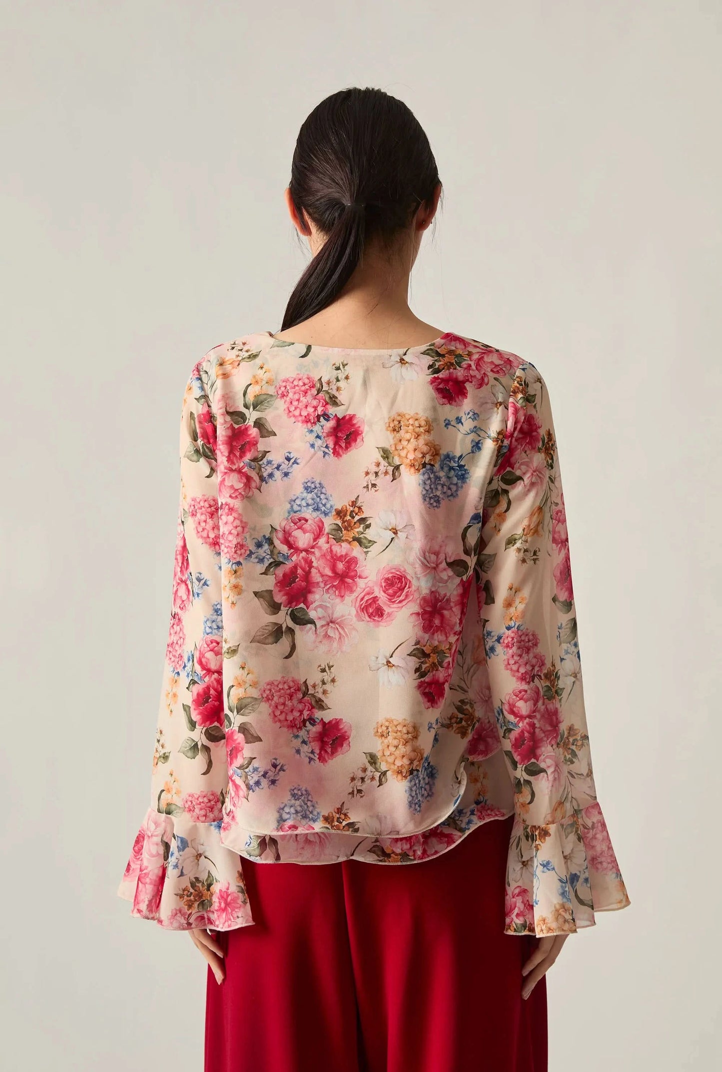 Back view of a woman wearing a floral blouse with red wide‑leg trousers against a soft beige background.
