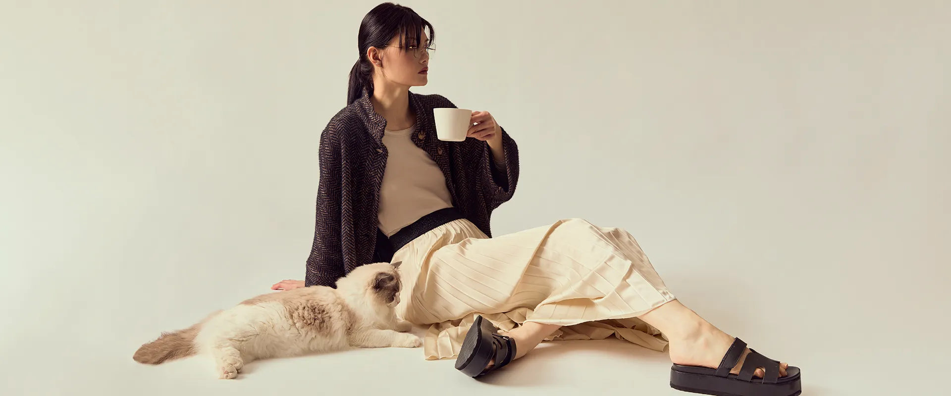 Woman sitting on the floor with a cat, holding a cup, wearing a black and purple jacket and beige skirt.