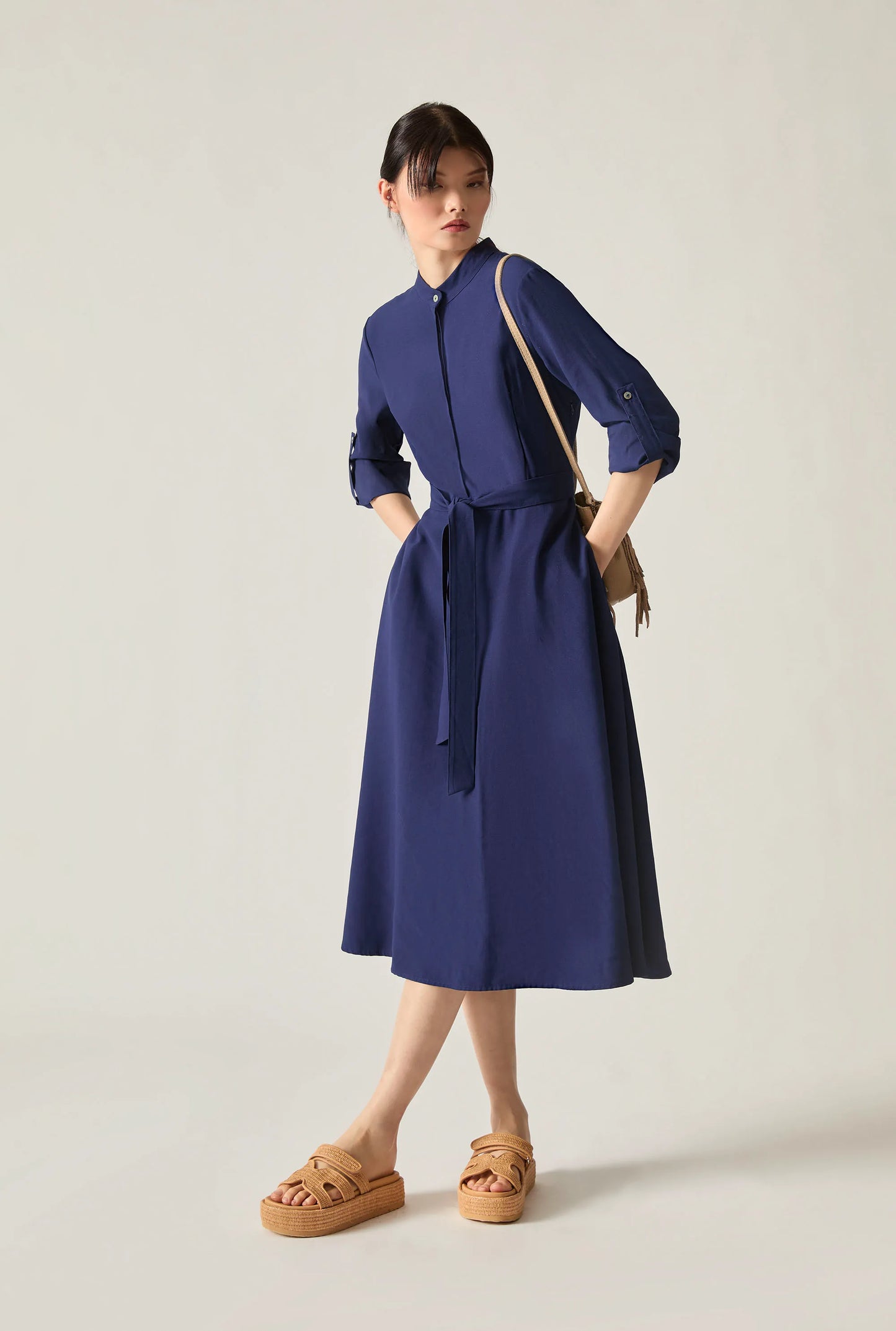 Woman wearing a navy tie‑waist midi dress with three‑quarter sleeves and tan platform sandals, standing against a soft beige background.