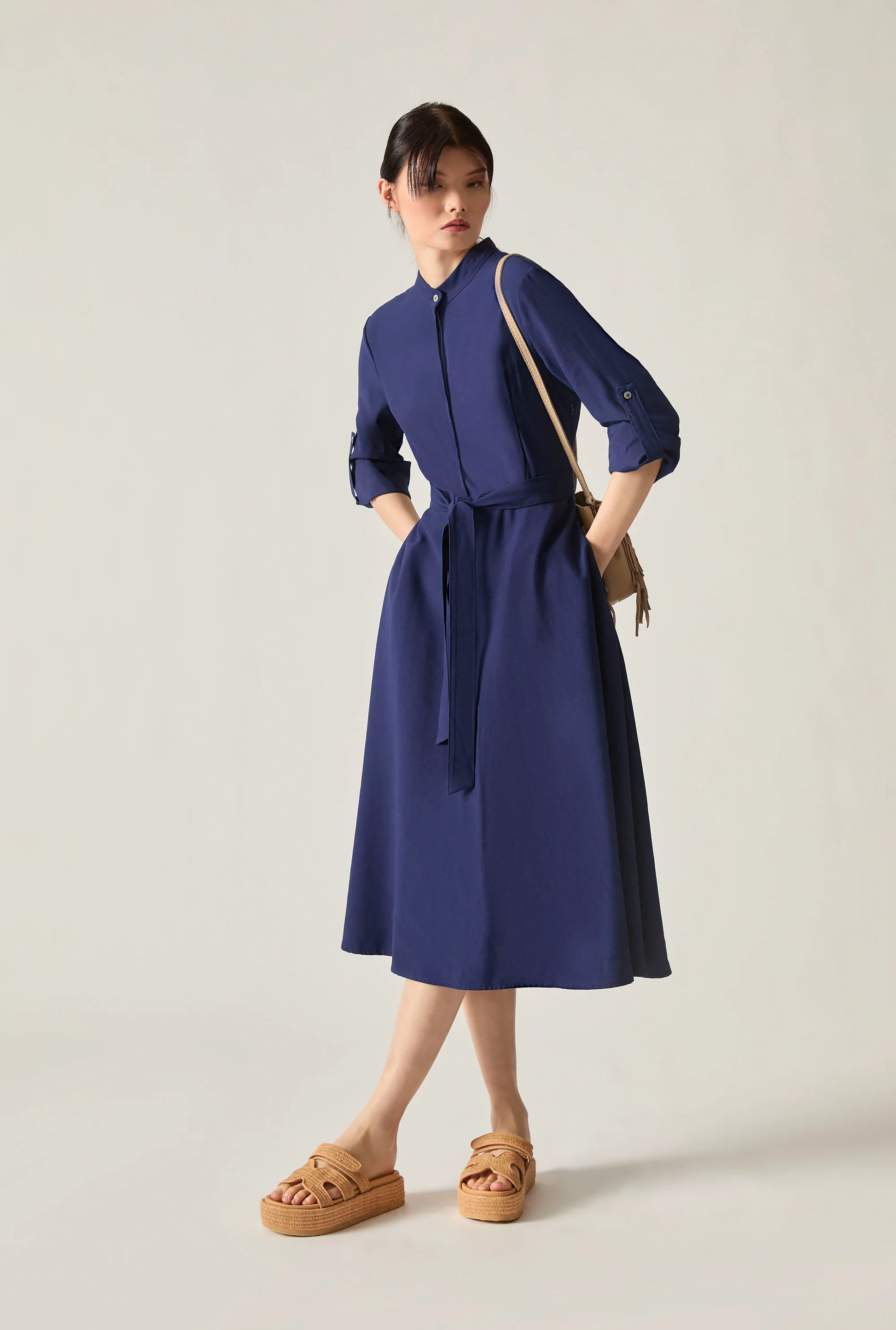 Woman wearing a navy tie‑waist midi dress with three‑quarter sleeves and tan platform sandals, standing against a soft beige background.