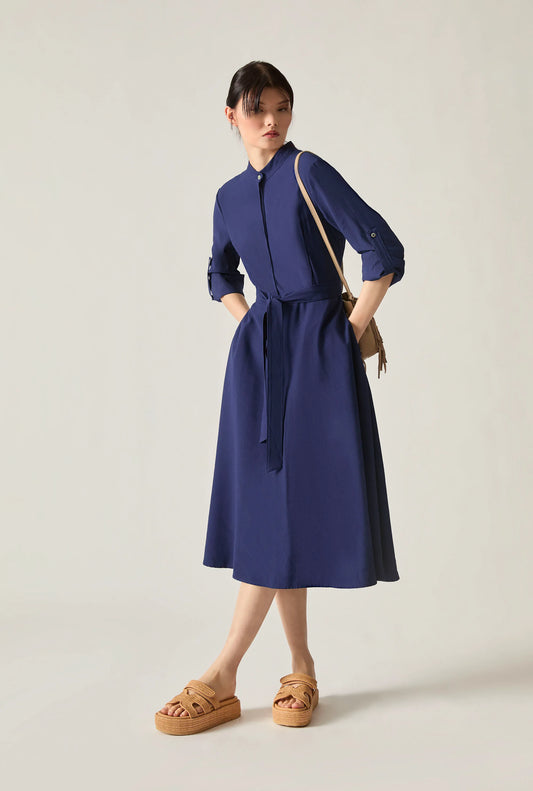 Woman wearing a navy tie‑waist midi dress with three‑quarter sleeves and tan platform sandals, standing against a soft beige background.