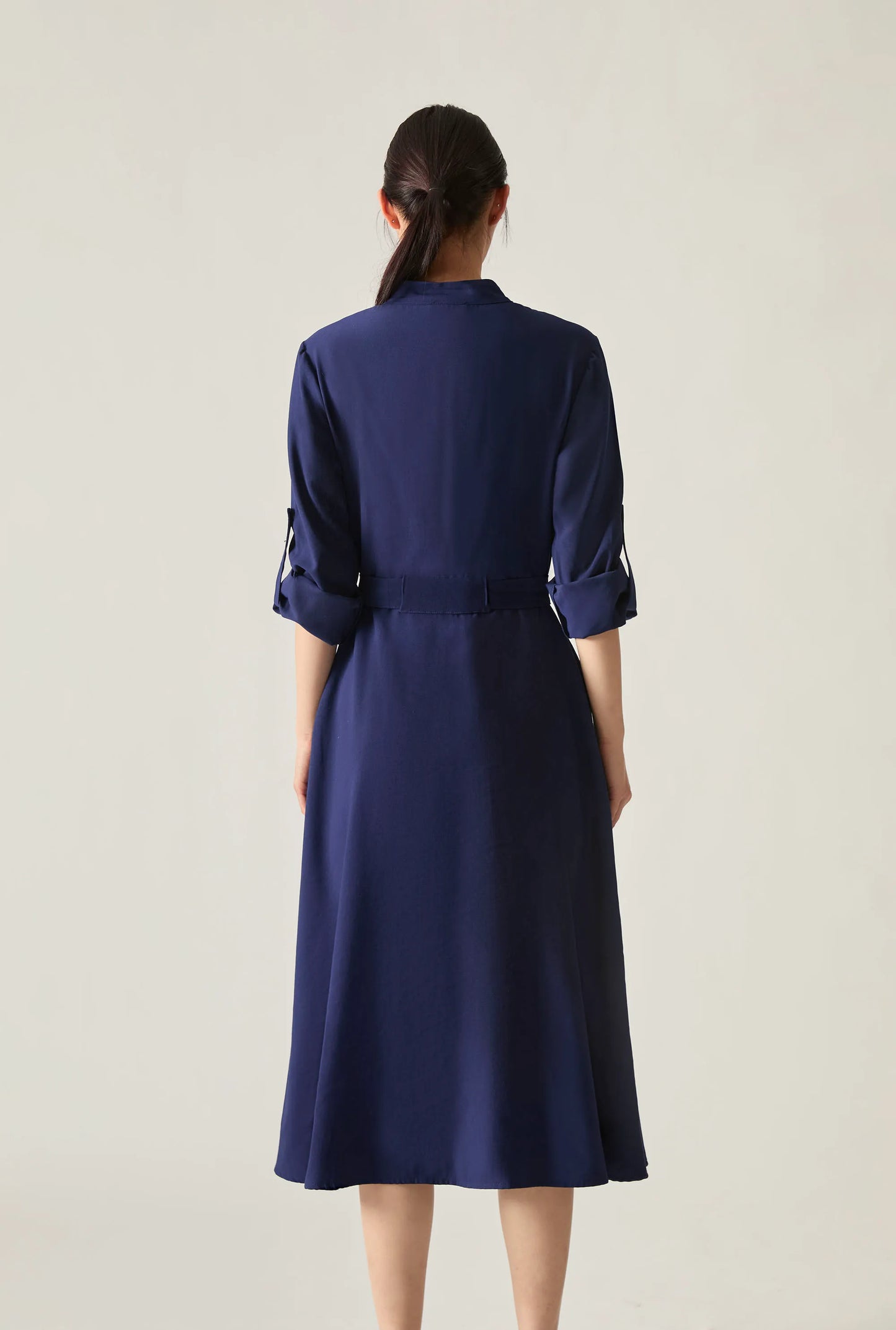 Back view of the navy tie‑waist midi dress with three‑quarter sleeves, shown against a soft beige background.